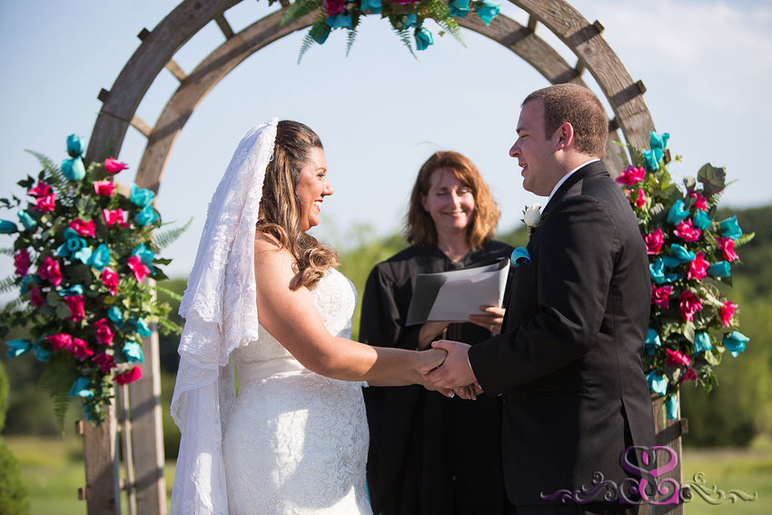 23-bride-and-groom-smile-during-ceremony-under-wood-arch-lawrence-photographer-stony-point-hall-wedding